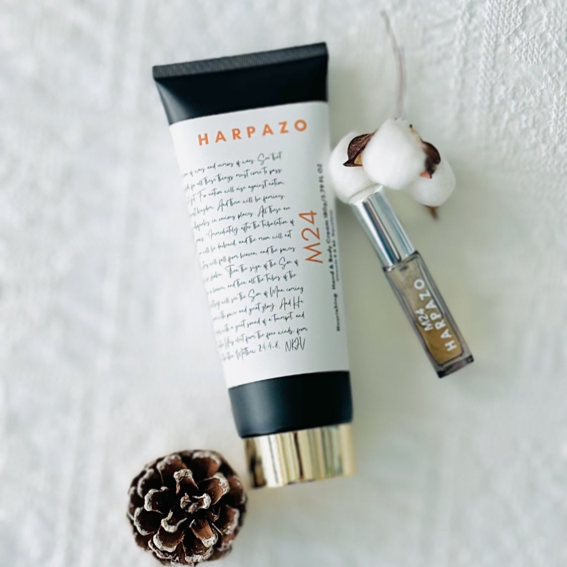 A tube of M24 hand and body cream with a capacity of 180g, accompanied by a travel perfume bottle and a cotton flower, presented on a white surface with a pine cone.