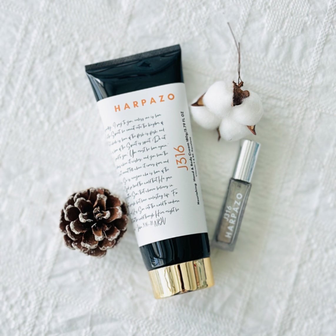 A tube of J316 Hand & Body Cream next to a small spray bottle of perfume, decorative cotton, and a pine cone on a white textured surface.
