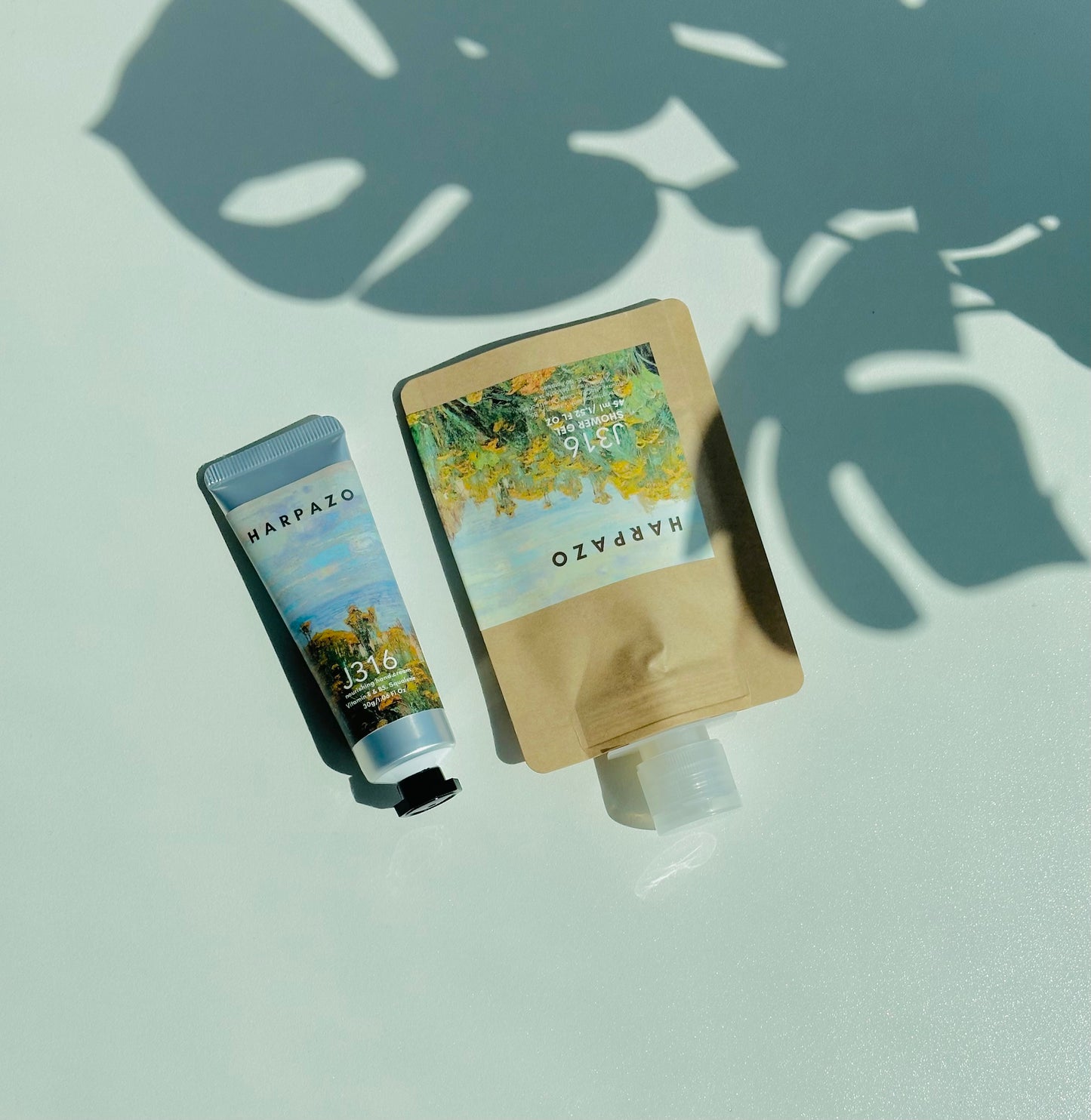 A travel care set consisting of a moisturizing hand cream tube and a travel shower gel bottle, displayed on a light background with a shadow of a plant leaf across the surface.