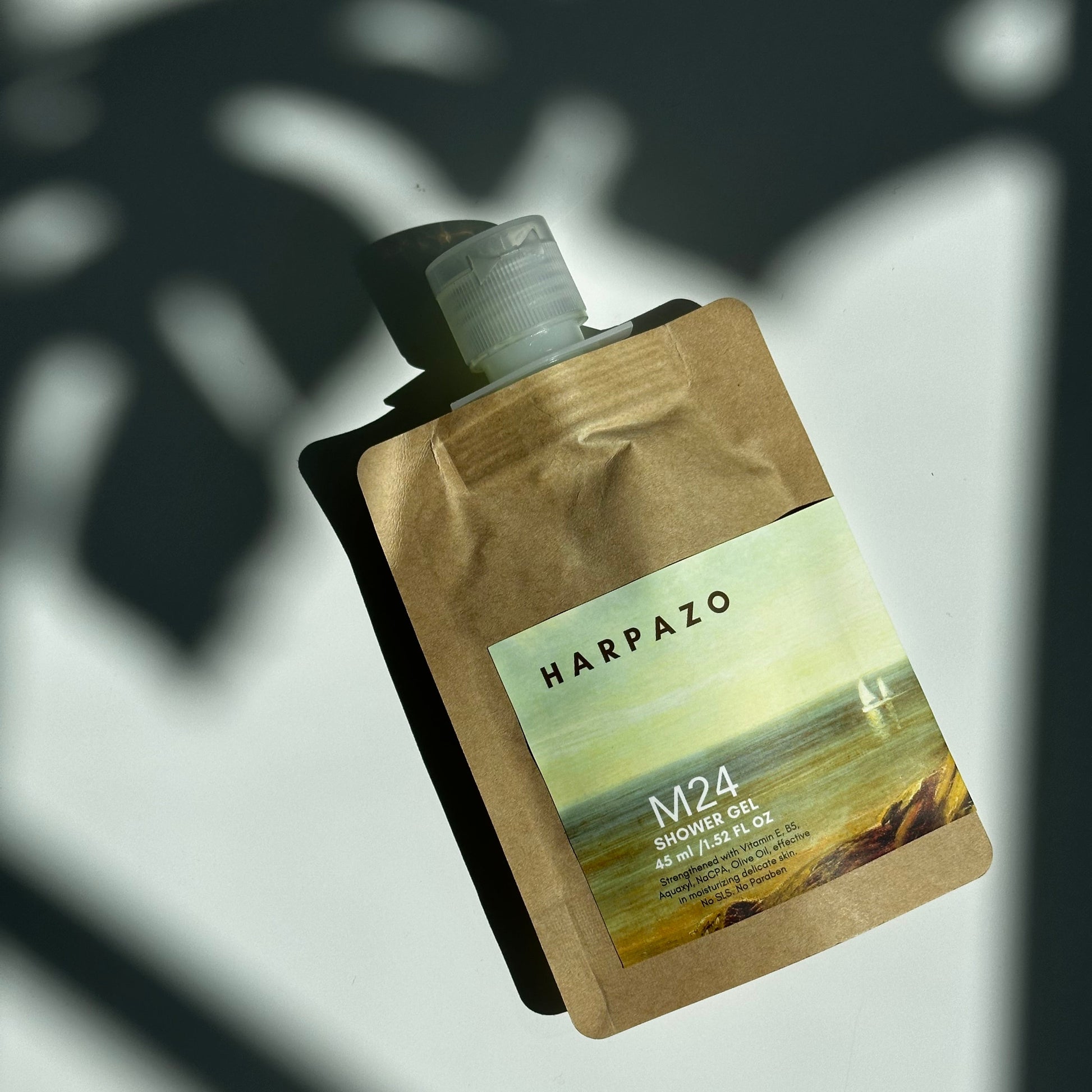 Travel size shower gel bottle with a beige to brown gradient packaging, labeled 'HARPAZO M24', with an image of a green landscape and clouds.