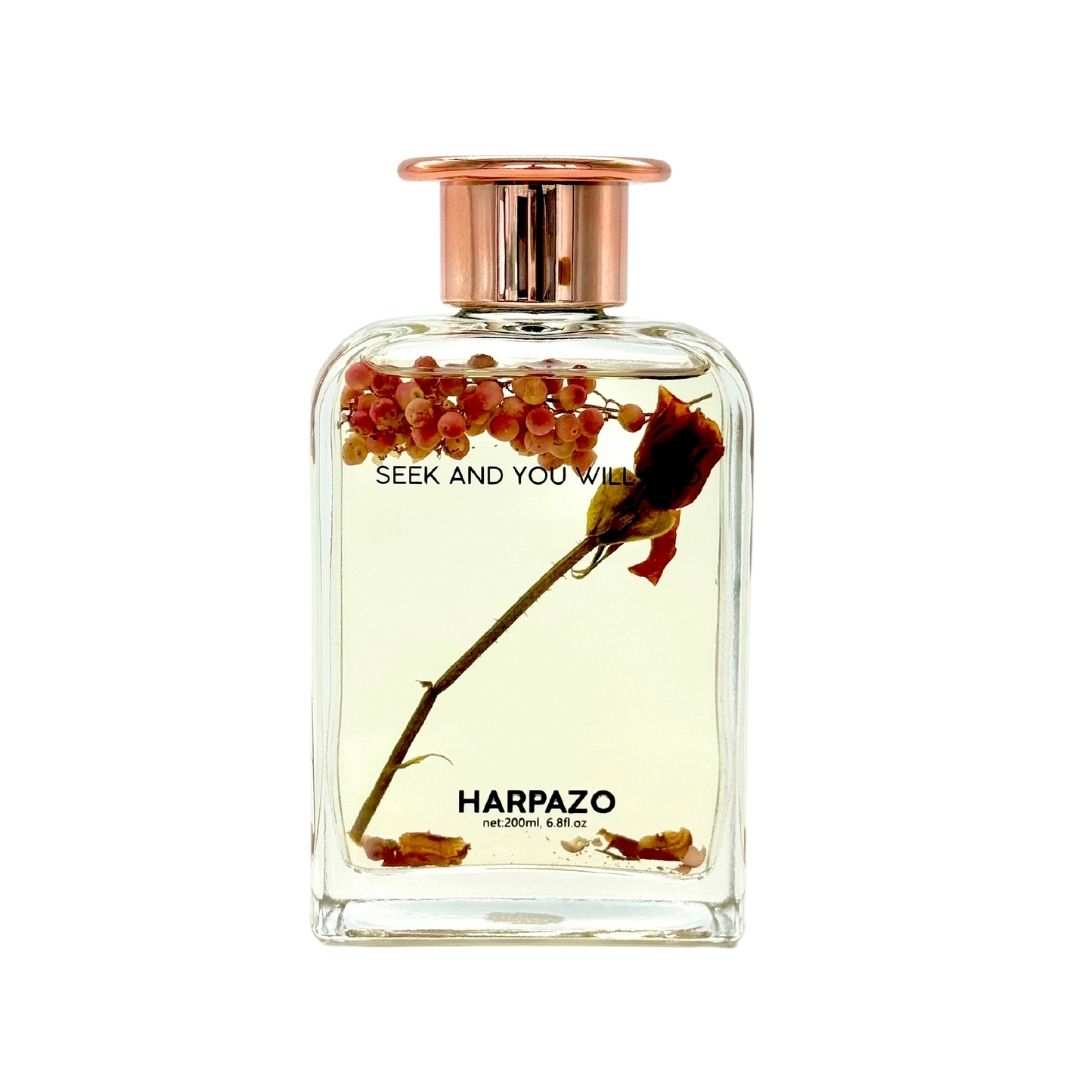 A clear glass reed diffuser bottle with a pink floral fragrance oil, branded with 'HARPAZO' and a phrase 'seek and you shall find'. The bottle is sealed with a rose gold cap.