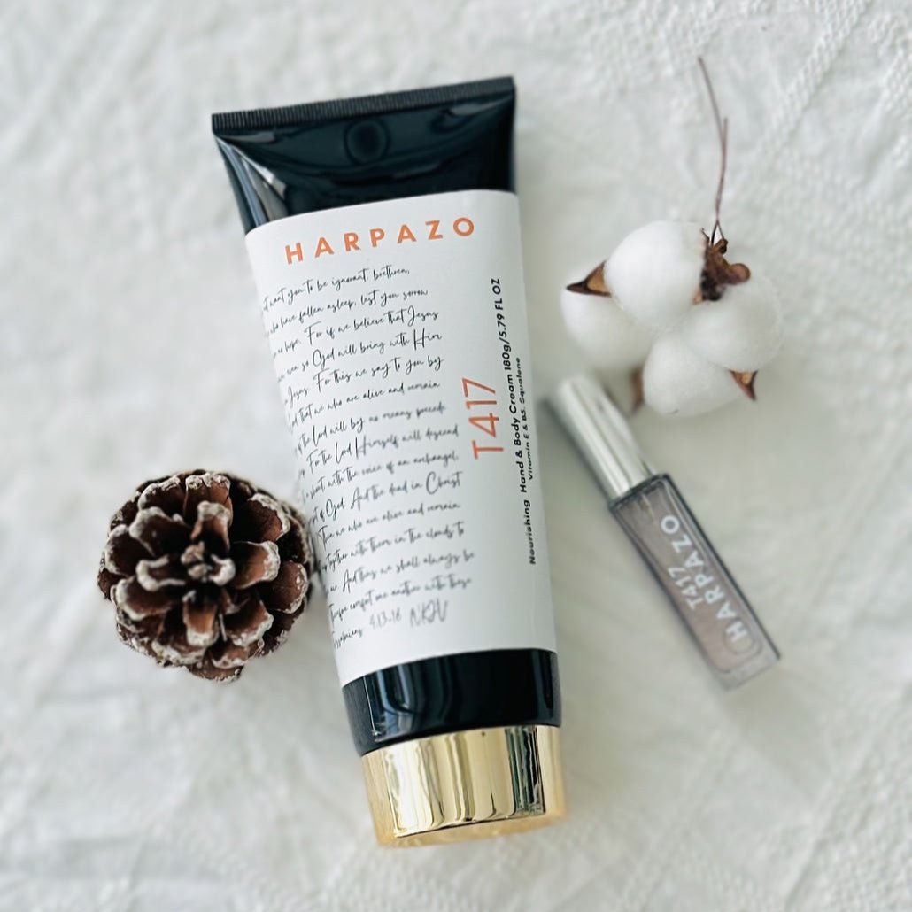 A tube of T417 hand and body cream with a gold-colored cap, placed next to a small perfume bottle and a pine cone, on a white textured surface.
