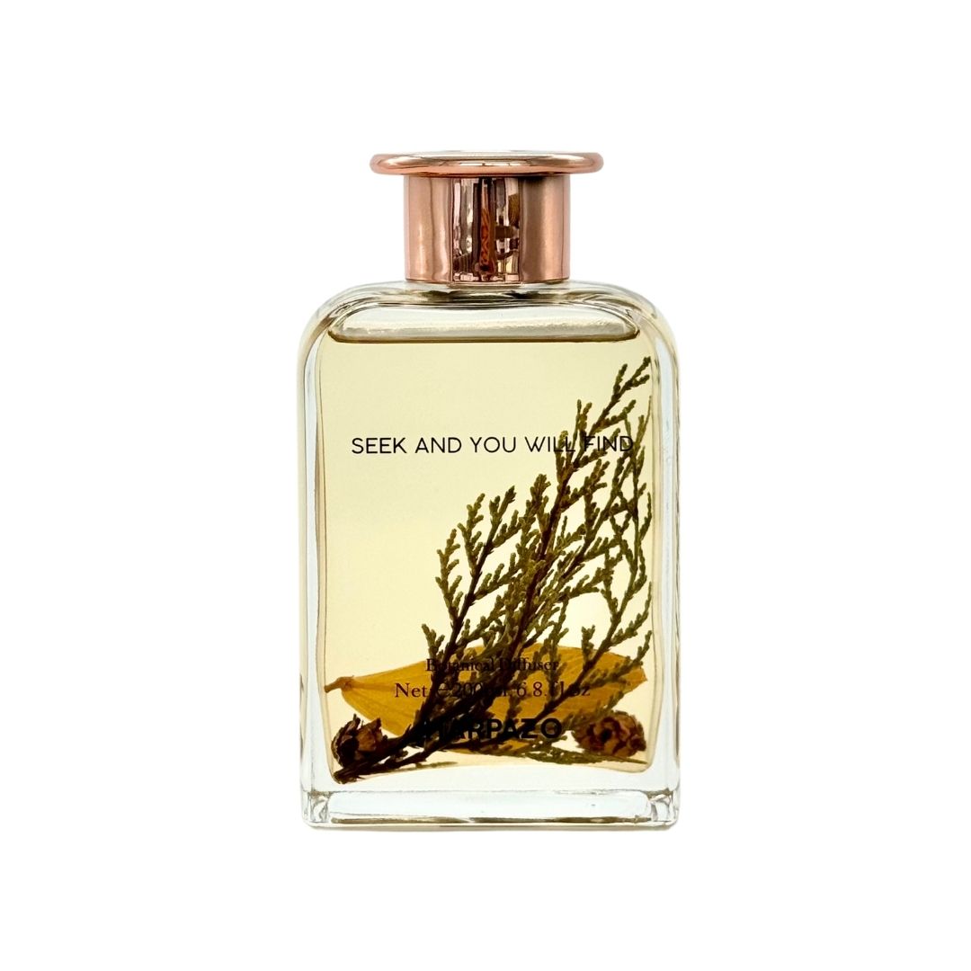 A clear glass reed diffuser bottle with a copper-colored cap and Longjing green tea leaves inside.