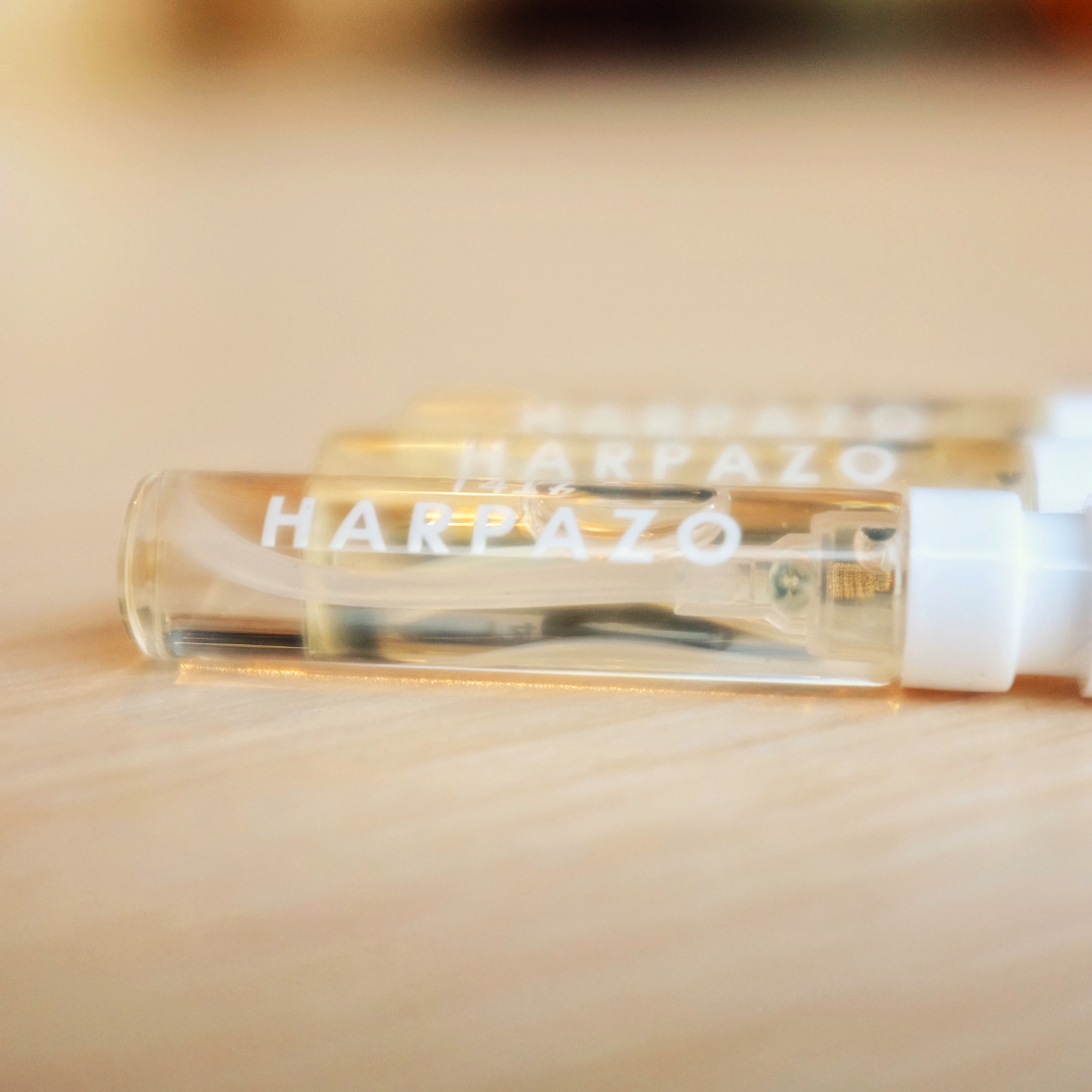 Three small, clear glass vials with white caps labeled with the brand 'Harpazo', containing perfume, arranged on a wooden surface.