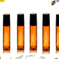 A set of six orange glass bottles with black caps, labeled with different names, arranged in a row against a white background with dried flowers around them.