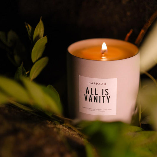 A white candle labeled 'ALL IS VANITY' with a lit flame, placed among green leaves.