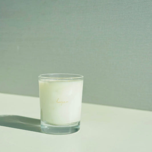 A white aromatic candle in a clear glass container, labeled 'Harpazo, placed on a light-colored surface with a neutral background.