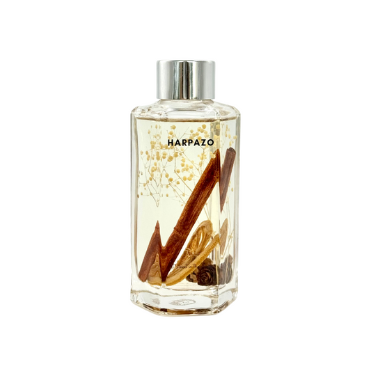 A clear glass reed diffuser bottle with a silver cap, filled with a brownish fragrance oil and several natural plants submerged within.