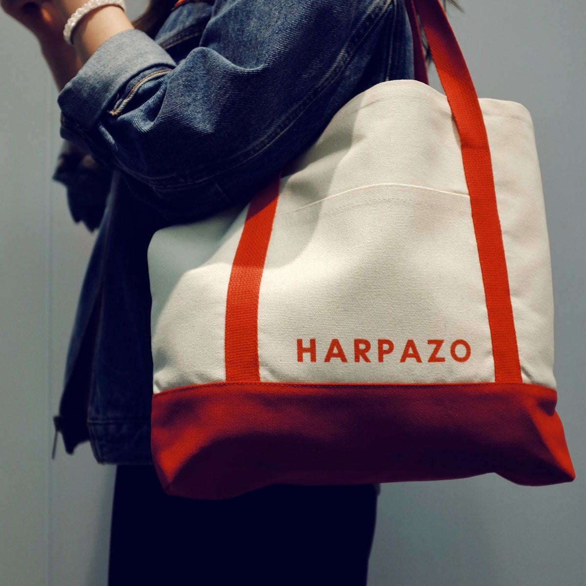 A person holding a HARPAZO travel tote bag in white with red stripes and the brand name 'HARPAZO' printed in red on the side.