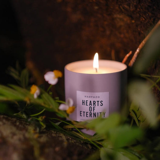 A purple candle labeled 'Hearts of Eternity' burning outdoors surrounded by greenery and a few flowers visible.