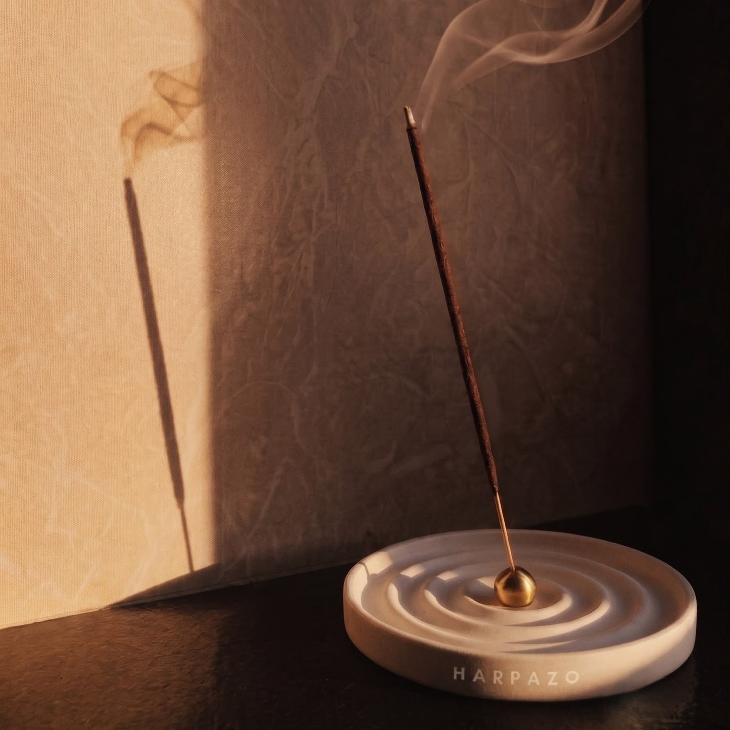 A HARPZO incense holder with a single burning incense stick, casting a shadow on a textured beige wall under dramatic warm lighting.