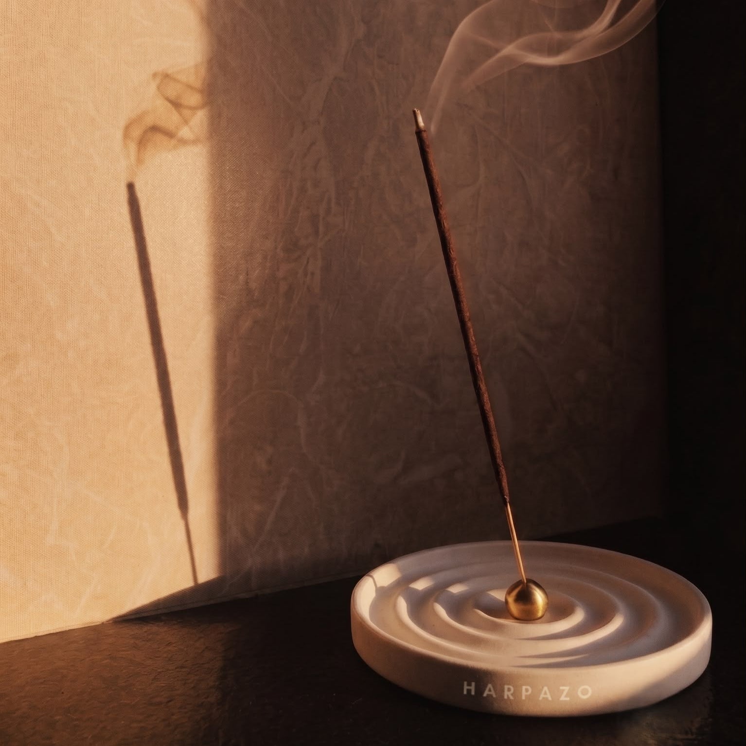 A HARPZO incense holder with a single burning incense stick, casting a shadow on a textured beige wall under dramatic warm lighting.