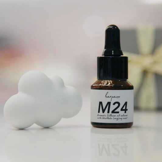 A bottle of M24 aromatic diffuser oil next to a white diffusing cloud on a reflective surface, with a blurred background.
