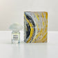 A small clear glass perfume bottle with a frosted cloud-shaped cap stands beside a tall rectangular box featuring an abstract swirl design in yellow, black, white, and gray. The label on both the bottle and box reads MARAZZO and miracle.