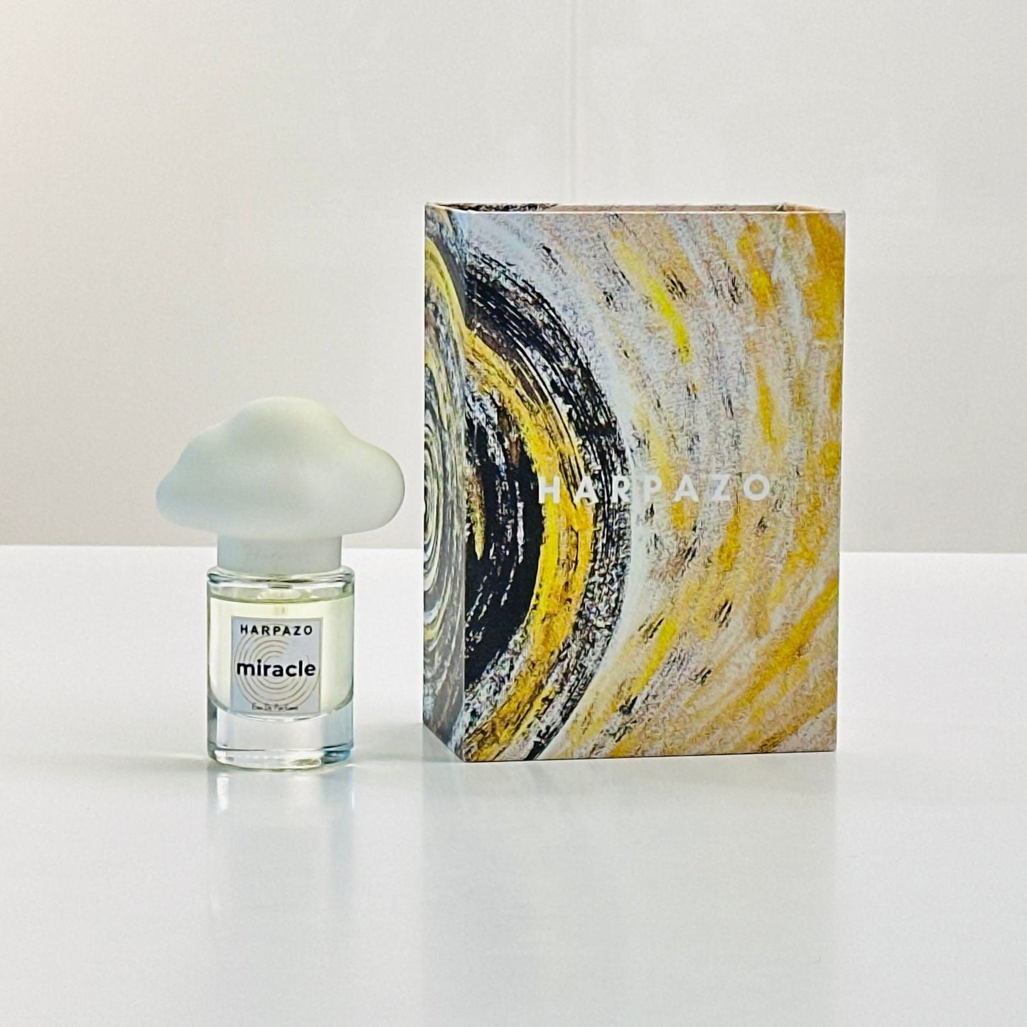 A small clear glass perfume bottle with a frosted cloud-shaped cap stands beside a tall rectangular box featuring an abstract swirl design in yellow, black, white, and gray. The label on both the bottle and box reads MARAZZO and miracle.