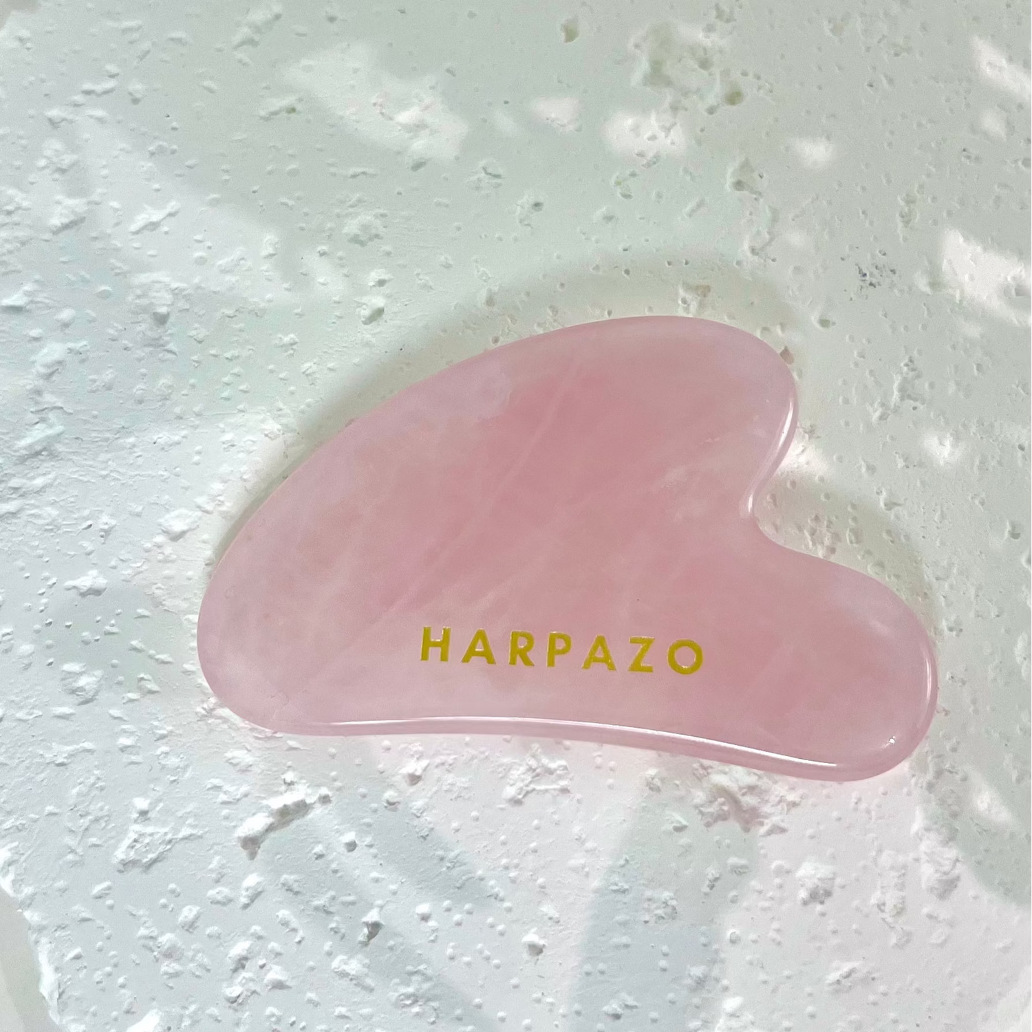 Pink gua sha tool with 'Harpazo' branding on a textured white background
