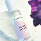 Harpazo Rose Mist bottle on a textured surface with a blurred purple flower in the background