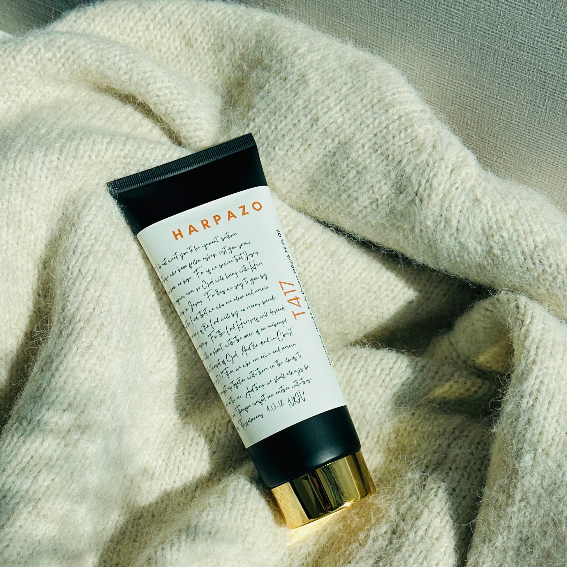 A tube of T417 Body and Hand Cream is lying on a beige fabric surface. The packaging is predominantly white with black and orange text, and the tube cap is gold-colored.