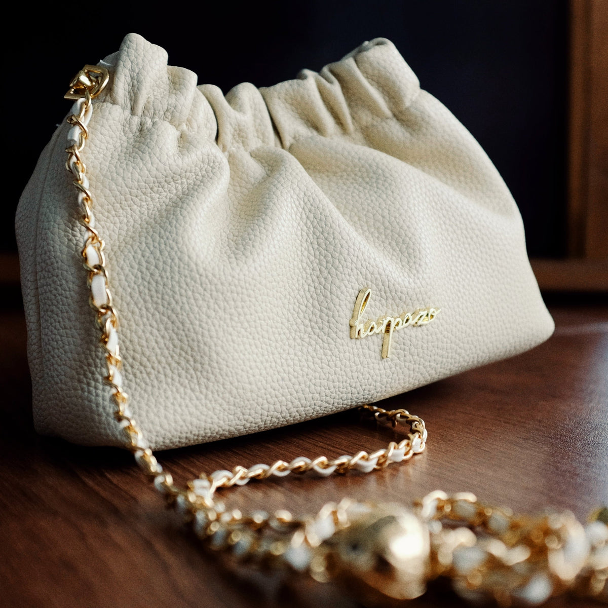 A small, beige handbag with a lychee texture and a gold chain strap, displayed against a wooden background.