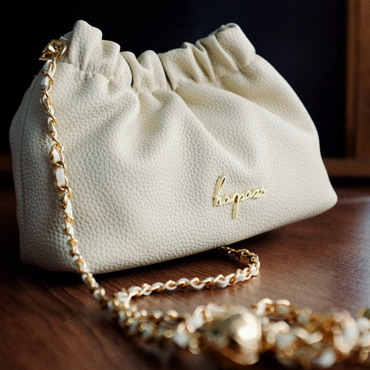 A small, beige handbag with a lychee texture and a gold chain strap, displayed against a wooden background.