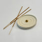 A round textured ceramic incense holder with concentric ripple patterns, featuring a small gold bead in the center, with three incense sticks resting on top, set against a clean light gray background.