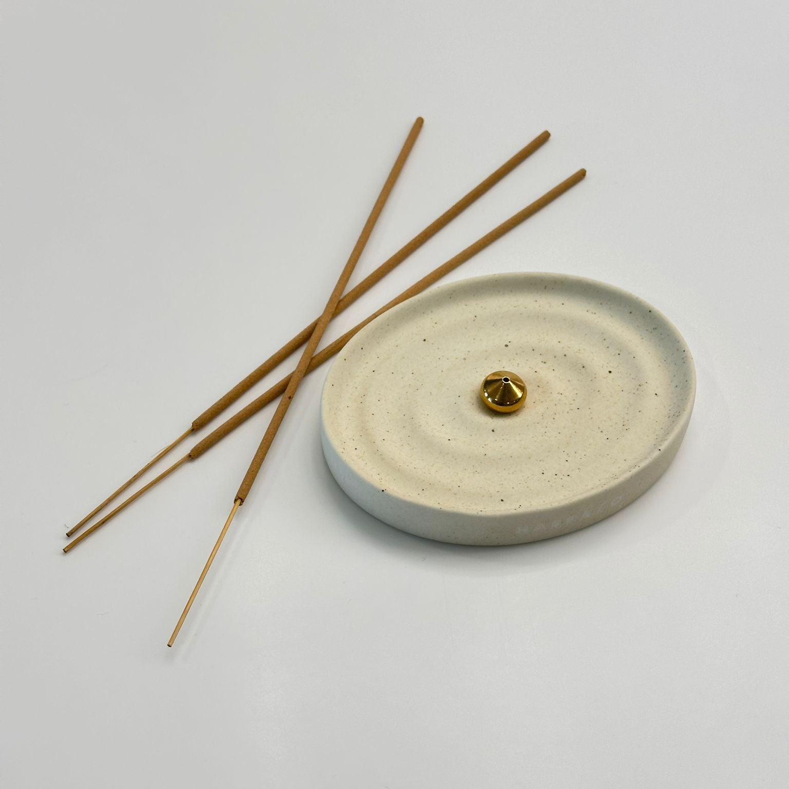 A round textured ceramic incense holder with concentric ripple patterns, featuring a small gold bead in the center, with three incense sticks resting on top, set against a clean light gray background.