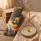 A HARPZO incense stick package labeled Mandarin & Basil lies on a rustic wooden table beside a lit incense stick in a round ceramic holder, with stacked books and draped fabric in the background.